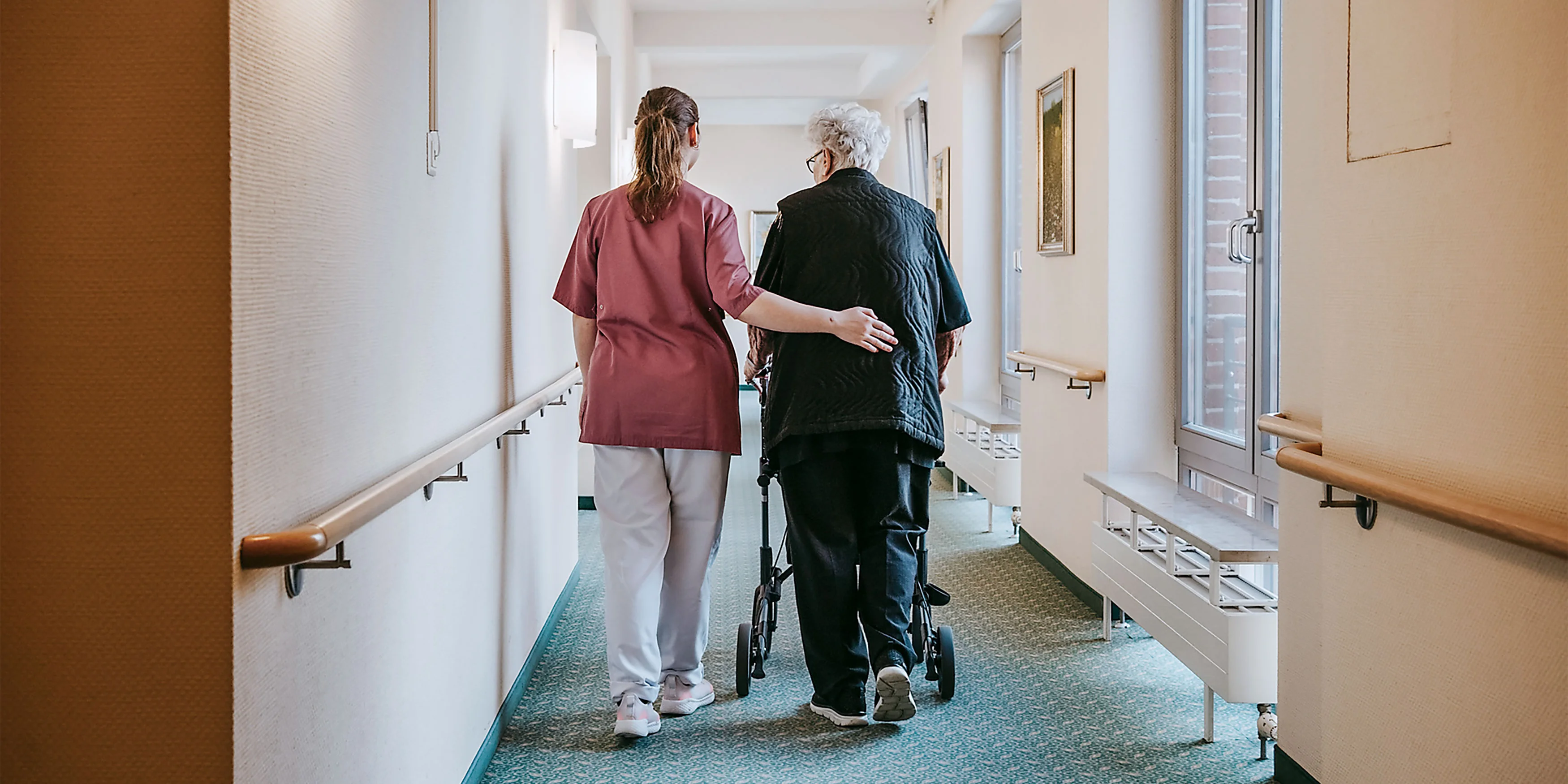 Pflegekraft stützt ältere Person mit Rollator beim Gehen durch einen hellen Flur in einer Einrichtung