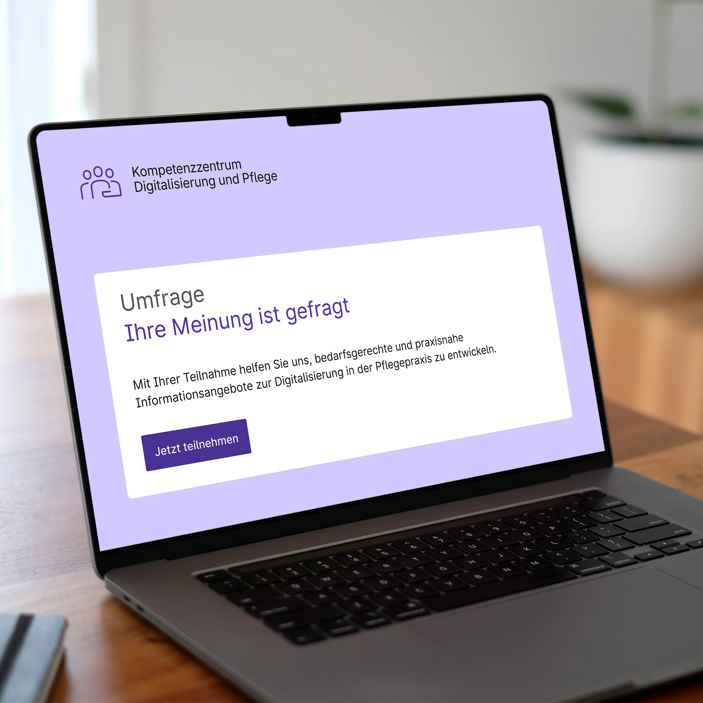 Aufgeklappter Laptop auf einem Holztisch. Im Display sieht man einen stilisierten Startbildschirm einer Umfrage des Kompetenzzentrums Digitalisierung und Pflege
