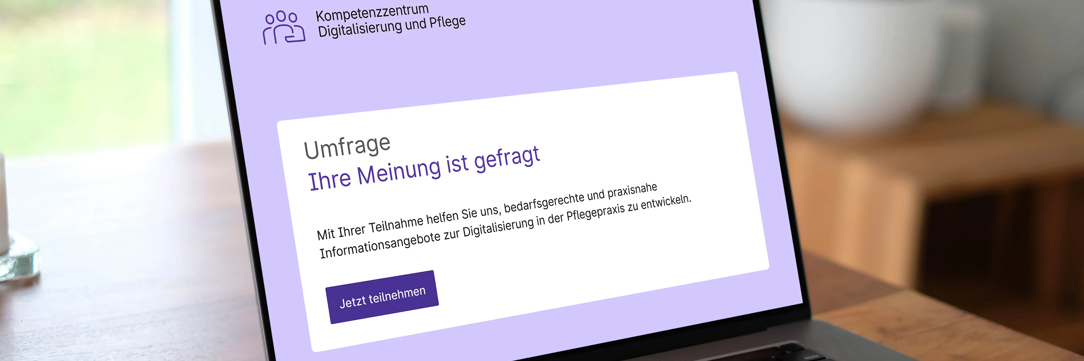 Aufgeklappter Laptop auf einem Holztisch. Im Display sieht man einen stilisierten Startbildschirm einer Umfrage des Kompetenzzentrums Digitalisierung und Pflege