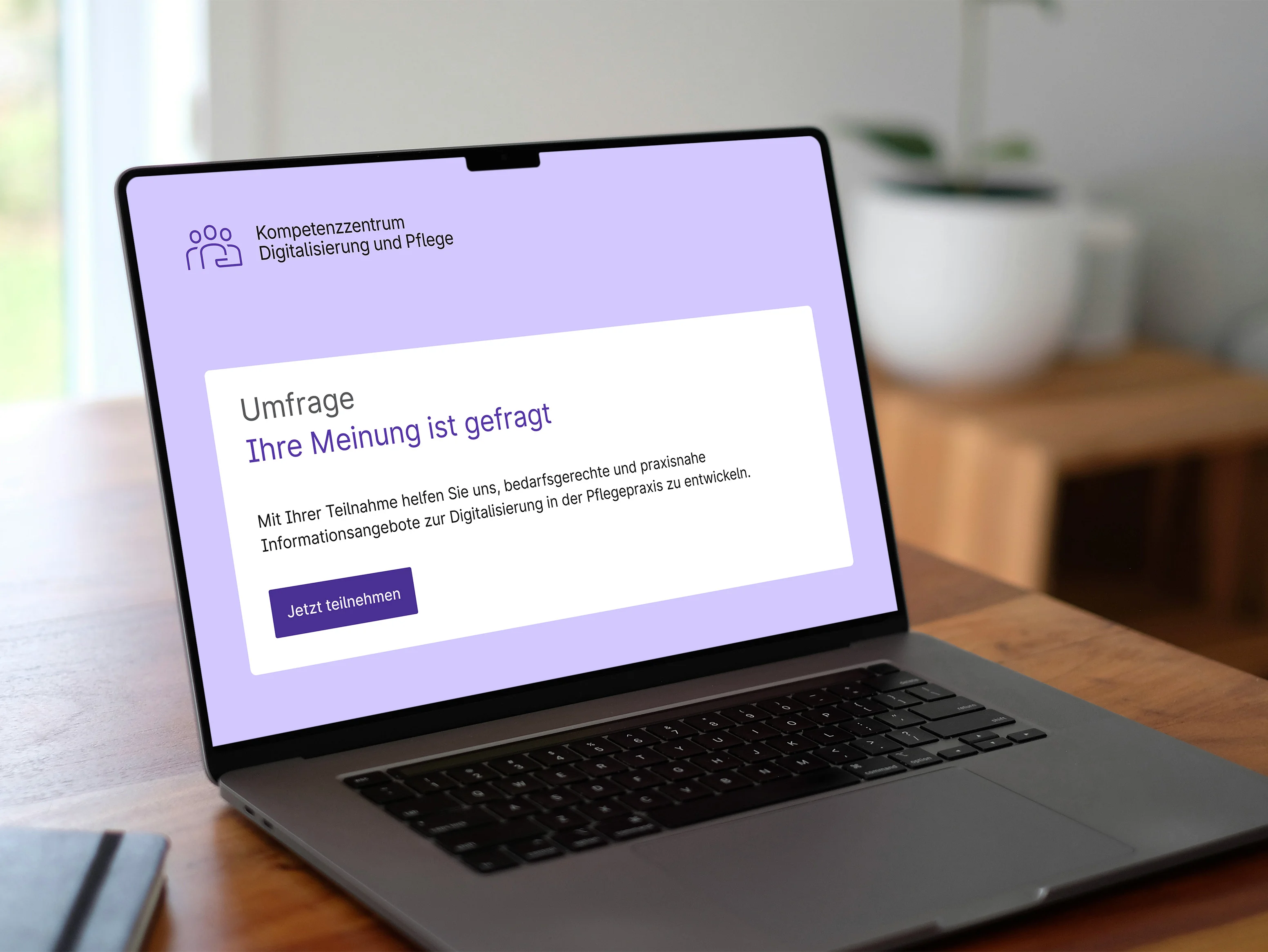 Aufgeklappter Laptop auf einem Holztisch. Im Display sieht man einen stilisierten Startbildschirm einer Umfrage des Kompetenzzentrums Digitalisierung und Pflege