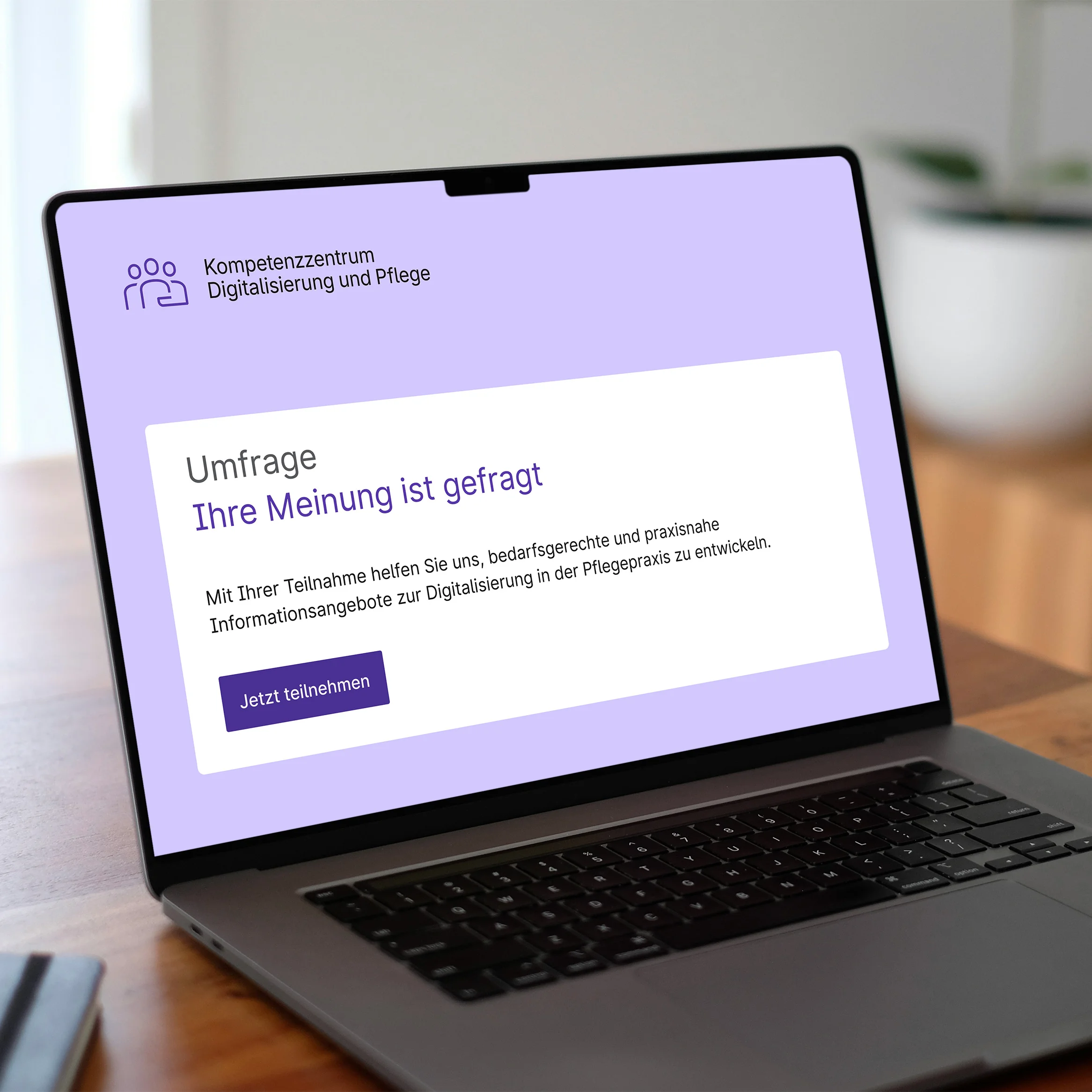 Aufgeklappter Laptop auf einem Holztisch. Im Display sieht man einen stilisierten Startbildschirm einer Umfrage des Kompetenzzentrums Digitalisierung und Pflege