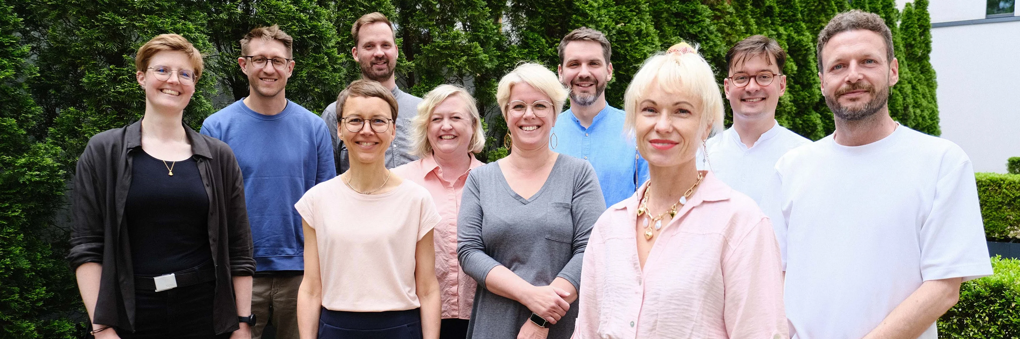 Gruppenfoto des Teams des Kompetenzzentrums Digitalisierung und Pflege im Freien vor grünen Hecken und einem modernen Gebäude.