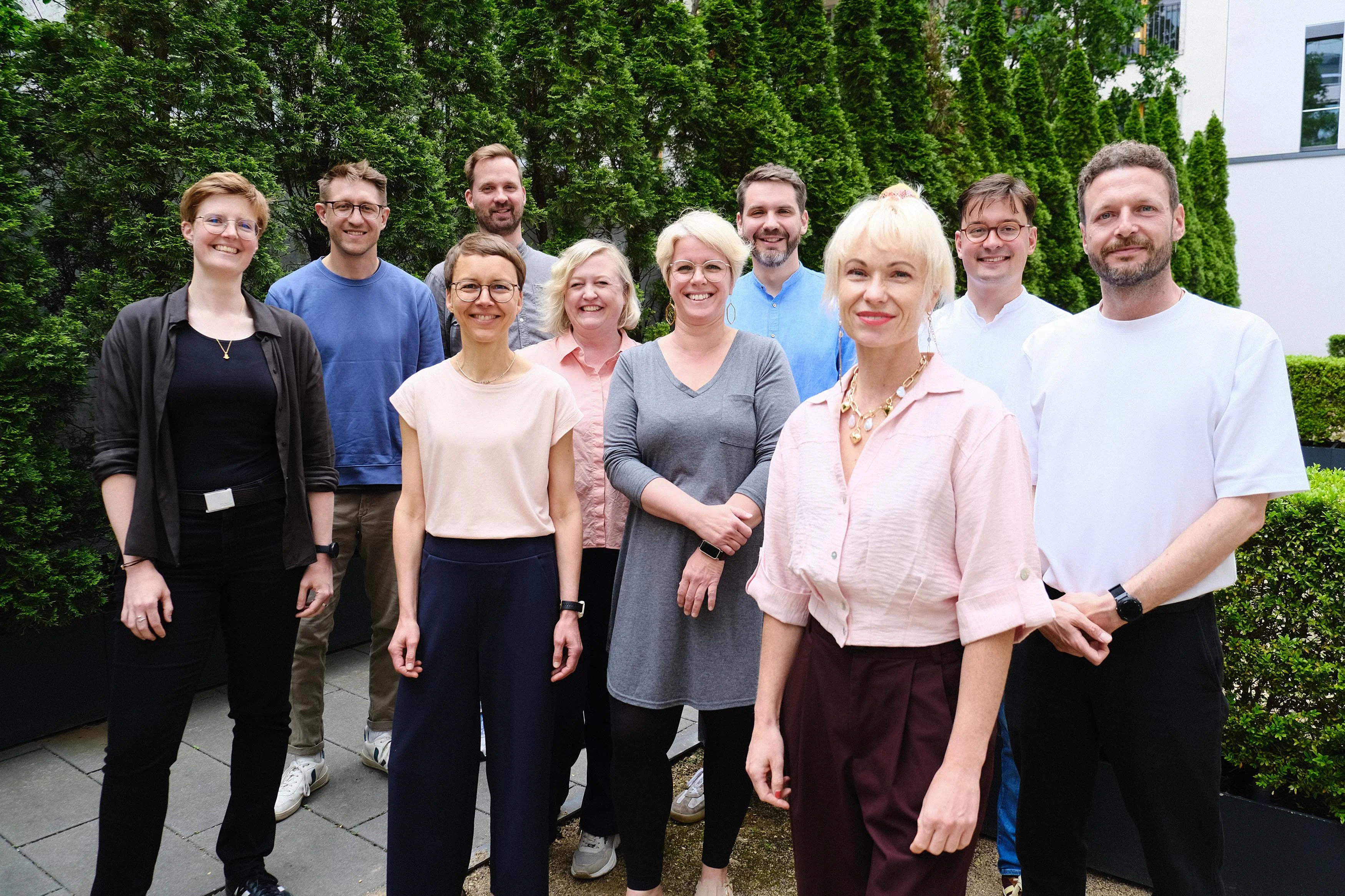 Gruppenfoto des Teams des Kompetenzzentrums Digitalisierung und Pflege im Freien vor grünen Hecken und einem modernen Gebäude.
