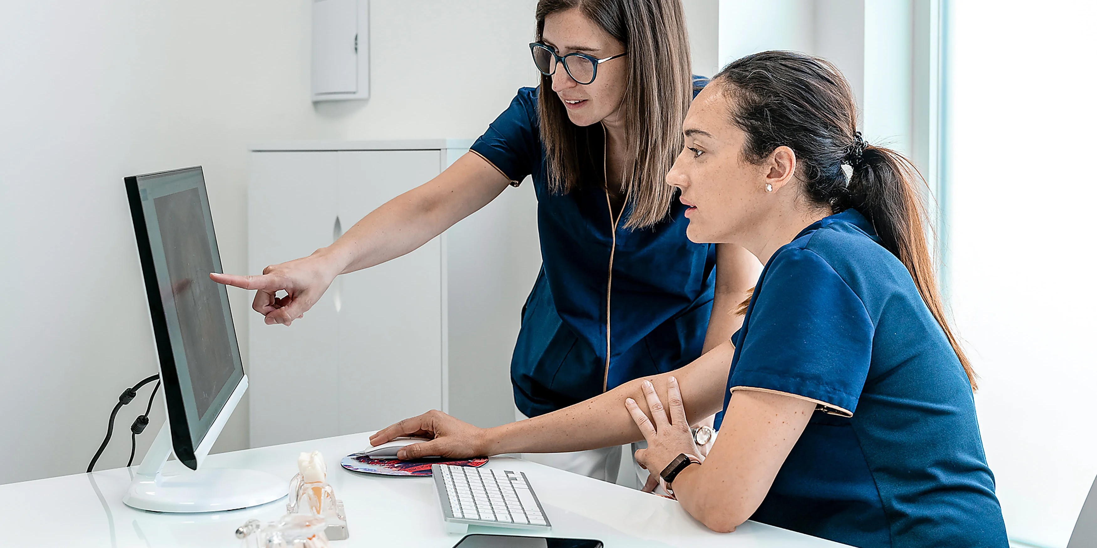 Zwei Pflegefachkräfte arbeiten an einem Computer; eine zeigt auf den Bildschirm, die andere bedient Tastatur und Maus.